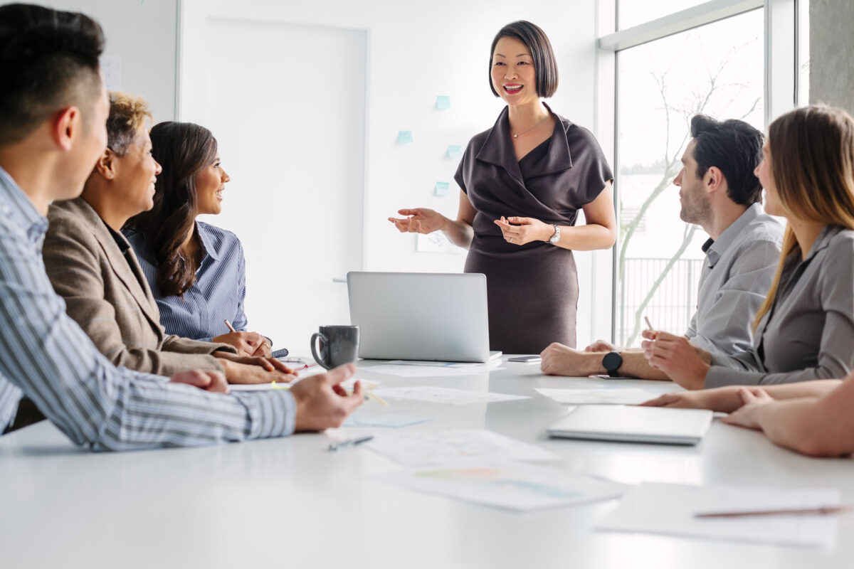 Korean mature woman leads meeting of multiracial work colleagues discussing ideas and business together sitting at table in modern creative office interior daytime
