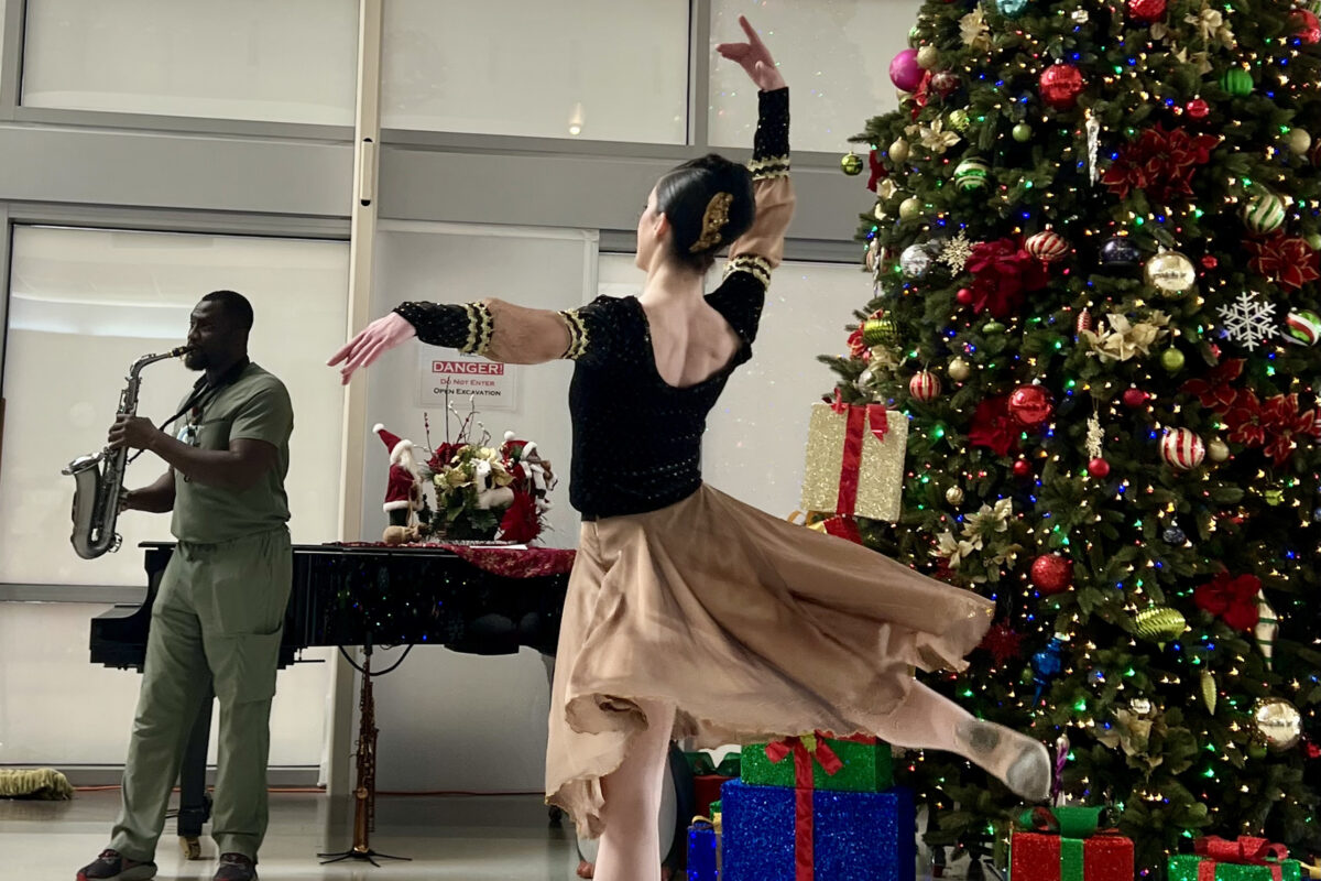 Nurse playing the saxophone while a ballerina dances in front of a Christmas tree.